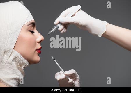 Seitenansicht des Patienten mit elastischem Verband in der Nähe von Ärzten, die ein Operationsmesser und eine Spritze isoliert auf dunkelgrau halten Stockfoto