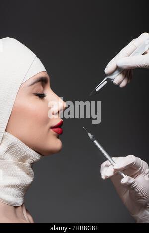 Seitenansicht einer Frau mit elastischem Verband auf dem Kopf in der Nähe von plastischen Chirurgen, die ein Operationsmesser und eine Spritze isoliert auf dunkelgrau halten Stockfoto