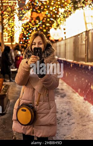 Eine junge Frau auf der Straße, auf dem Platz mit einem Weihnachtsbaum und Weihnachtsdekorationen. Neujahr und Weihnachten feiern Stockfoto