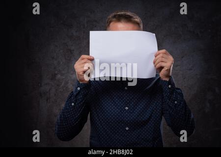 Ein junger Europäer hält ein weißes Blatt Papier in seinen Händen, das sein Gesicht bedeckt. Für Werbung auf dunklem Hintergrund Stockfoto