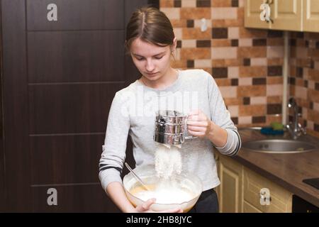 Das Mädchen gießt Mehl in den Teig. Das Mädchen kocht. Hände, die den Teig zubereiten. Frauen kneten frischen Teig für die Herstellung Stockfoto