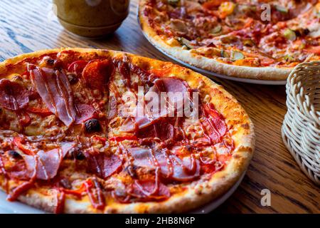 Zwei frisch gebackene, hausgemachte, in Scheiben geschnittene Pizzas mit Käse- und Schinkenaufschnitt, serviert mit Geschirr auf Holzhintergrund. Stockfoto