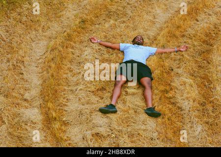 Schwarzer Mann legt sich im Feld nieder. Junger Teenager im Feld. Mann im Weizenfeld. Junge Person auf dem Land. Afroamerikanischer schwarzer Mann liegt im Weizenfeld. Stockfoto