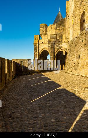 Ein doppeltes spitzbogiges Tor führt durch die Ringmauer der Cité de Carcassonne, Département Aude, Frankreich, auf einen Kopfsteinpflasterweg, der von langen Abendschatten überquert wird. Stockfoto