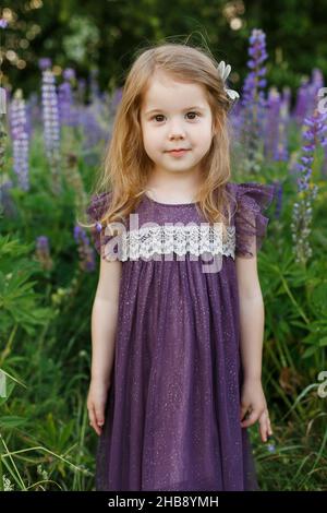 Nahaufnahme Porträt von niedlichen, vier Jahre alt, kleines Mädchen in violettem Kleid. Lächelndes Kind, das sich unter dem Feld der Lupinenblumen amüsieren kann. Kinder spielen draußen. Concep Stockfoto