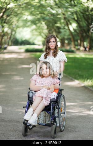 Attraktive Frau in Kleid mit Blumenmuster steht im grünen Park hinter ihrer Freundin, die Rollstuhl benutzt. Zwei Damen, die gemeinsam auf der Natur spazieren. Assistenz- und Supportkonzept. Stockfoto