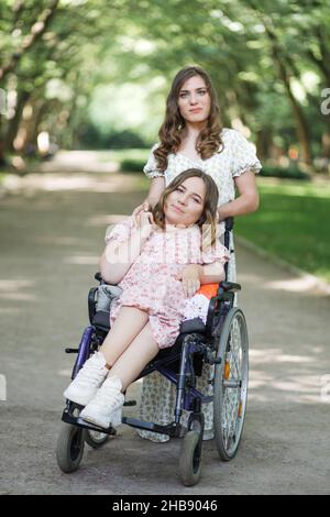 Attraktive Frau in Kleid mit Blumenmuster steht im grünen Park hinter ihrer Freundin, die Rollstuhl benutzt. Zwei Damen, die gemeinsam auf der Natur spazieren. Assistenz- und Supportkonzept. Stockfoto