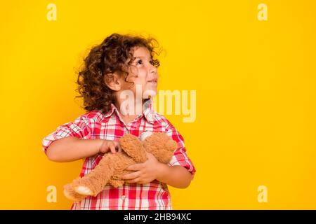Foto von attraktiven verträumten Schulmädchen gekleidet karierte Kleidung halten Spielzeug suchen leeren Raum lächelnd isoliert gelbe Farbe Hintergrund Stockfoto