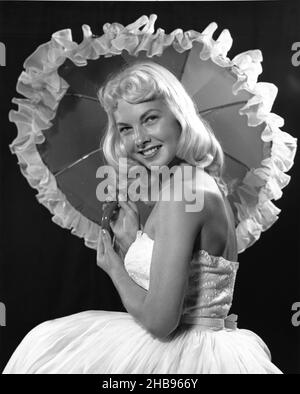 Blonde Mädchen in weißem Hochzeitskleid und Regenschirm mit Rüschen um die Kanten posiert, während Blick auf die Kamera. Ca. 1965 Stockfoto