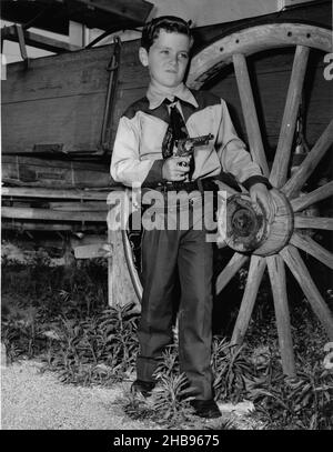 Kleiner Junge im Cowboy-Outfit mit einer Spielzeugpistole, die neben einer Postkutsche steht, um 1955 Stockfoto