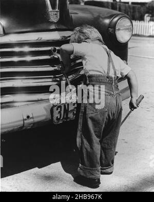 Kleiner Junge in Overalls mit Schraubendreher und Schraubenschlüssel, um zu handeln, als würde er ein Auto reparieren. Ca. 1945 Stockfoto