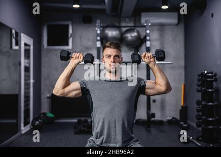 Muskulöser Kerl, der Hantel hebt, während er auf einer Bank im Fitnessstudio sitzt. Ein junger Athlet, der während eines Trainings Kurzhanteln verwendet. Starker Mann unter körperlicher Anstrengung Stockfoto