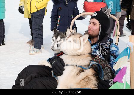 Dudinka, Region Krasnojarsk, Russland, 13. April 2019: Ein junger Mann in warmen, hellen Kleidern sitzt in einem Schlitten und umarmt seinen Husky Dog. Festival des Eisfiles Stockfoto