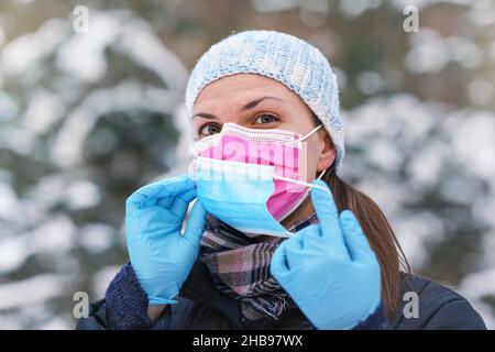 Junge Frau in warmer Winterkleidung trägt rosa Einweg-Virus Gesichtsmaske, setzen eine andere auf - einige raten, dass zwei Schichten bietet bessere Prot Stockfoto