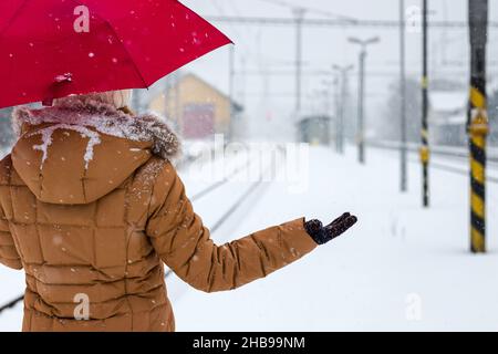 Wo ist der Zug? Verzögerter Zug während der Schneefälle. Frau mit rotem Regenschirm wartet im Winter beim Bahnhof auf Schneefall Stockfoto