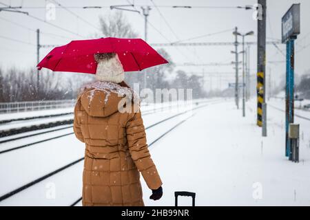 Frau, die während des Schnees am Bahnhof auf den Zug wartete, mit einem Regenschirm. Eisenbahn im Winter. Aktiv- und Reise-Lifestyle-Konzept Stockfoto