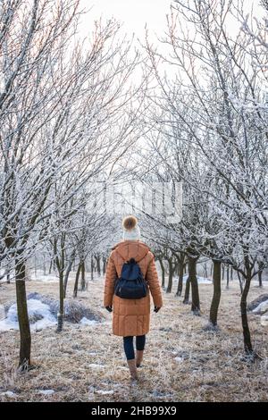 Frau in Winterkleidung und Rucksack. Weibliche Touristen, die bei kaltem Wetter in einer gefrorenen Baumallee spazieren gehen. Wandern in der Natur Stockfoto