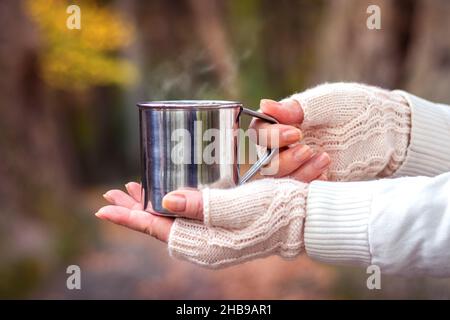 Frau mit gestricktem fingerlosen Handschuh hält den Becher mit dampfendem heißen Getränk. Erfrischung während der Wanderung in der Natur. Heißer Kaffee oder Tee in einer Metalltasse Stockfoto