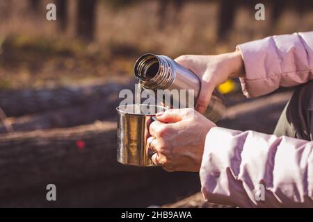 Frau gießt heißes Getränk aus Thermoskannen in einen Metallbecher im Herbstwald. Erfrischung beim Wandern in der Natur. Stockfoto