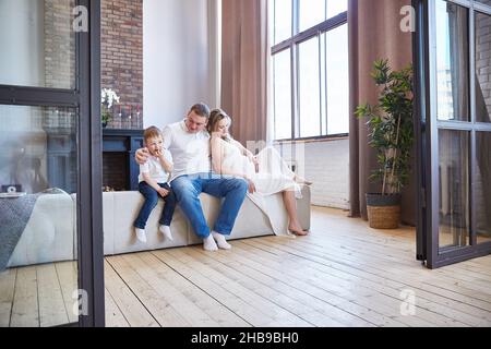 Eine glückliche Familie mit einem kleinen Kind und einer Schwangeren sitzt auf dem Sofa im Wohnzimmer. Speicherplatz kopieren Stockfoto