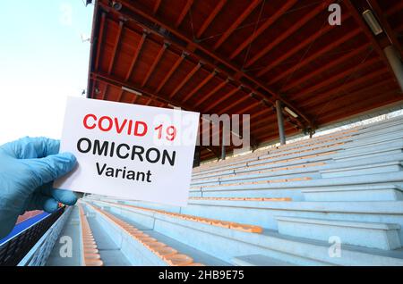 Covid 19 neue Omicron-Variante. Hand mit blauem Handschuh hält weiße Karte mit dem Text „Covid 19 Omicron Variant“ in einem Stadion mit leeren Tribünen. Conceptu Stockfoto