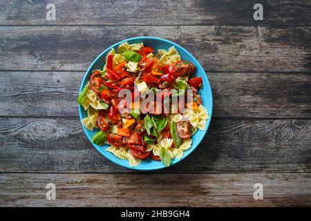Leichter und saftiger Sommersalat mit Nudeln, Tomaten, Käsewürfeln, Paprika und Basilikum in einer blauen Schüssel. Dunkler Holzhintergrund. Stockfoto