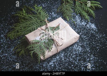 Ein Geschenk ist in Kraftpapier mit Tannenzweigen auf einem schwarzen Brett verpackt. Geschenkbox mit Schnee auf dunklem Hintergrund. Stockfoto