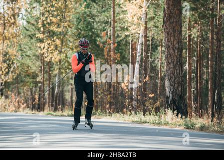 Training ein Athlet auf der Walze Skater. Biathlon Fahrt auf dem Roller Ski mit Skistöcke, im Helm. Herbst Training. Roller Sport. Stockfoto