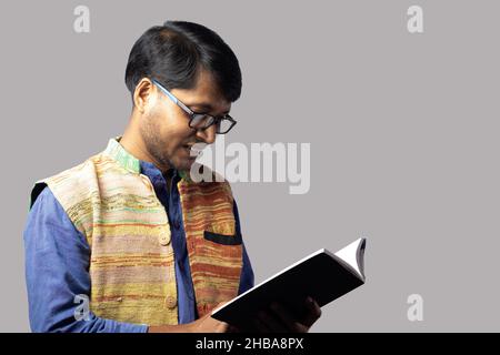 Ein junger indischer Mann in ethnischer Kleidung liest Buch auf grauem Hintergrund Stockfoto