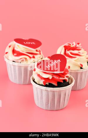Cupcake. Cupcakes in Herzform in weißen Tassen mit Inschriften: Liebe, glücklich auf rosa Hintergrund. Valentinstag. Vertikales Foto Stockfoto