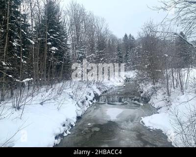 Winterlandschaft mit schneebedeckten Ästen. Der Bach, der in der Mitte fließt, ist teilweise gefroren und es gibt eine Schicht aus Schnee und Eis. Stockfoto