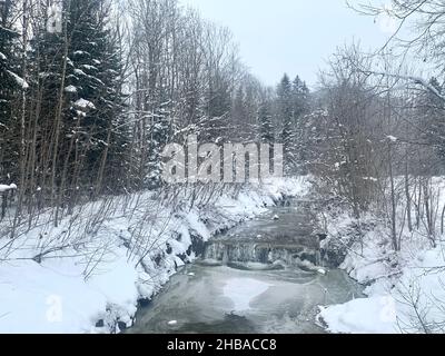 Winterlandschaft mit schneebedeckten Ästen. Der Bach, der in der Mitte fließt, ist teilweise gefroren und es gibt Schnee- und Eisschicht auf einer Kaskade. Stockfoto