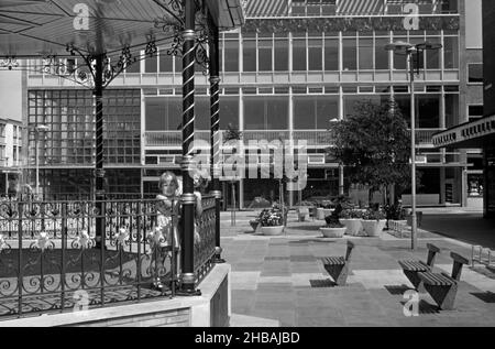 Queen’s Square, Crawley ‘New Town’, West Sussex, England, Großbritannien, mit zwei Mädchen, die auf dem legendären Bandstand spielen, der im Jahr 1960s fotografiert wurde. Die Erweiterung der wichtigsten Einkaufsmöglichkeiten war für Crawleys Expansion unerlässlich. Die erste Etappe, die eröffnet wurde, war der Broadwalk im Jahr 1954, nach der Eröffnung des Queen's Square-Bauvorbaus durch Ihre Majestät die Königin im Jahr 1958. Nach dem Zweiten Weltkrieg wurden viele Menschen und Arbeitsplätze in die neuen Städte im Südosten Englands verlegt, um diejenigen in Londons armen oder bombardierten Wohnungen zu verlagern, und Crawley war das einzige von ihnen – ein altes Foto aus dem Jahr 1960s. Stockfoto