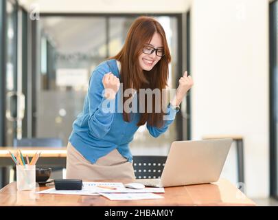 Junge Geschäftsfrau schaut mit hochgeklappten Händen auf den Laptop herunter und lächelt vor Freude, Mädchen in der Brille hebt ihre Hände vor Aufregung, Laptop Stockfoto