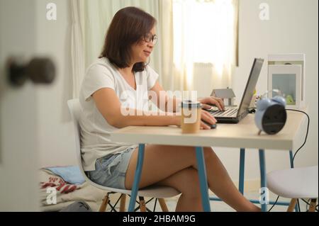 Eine Frau, die von zu Hause aus mit einem Laptop im Schlafzimmer arbeitet, ein weißes T-Shirt trägt, eine Brille trägt, ein lächelndes Gesicht trägt, die Hand mit der Maus und der Tastatur hält, d Stockfoto