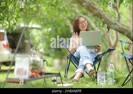 Eine Frau, die während des Campens mit einem Freund auf einem Laptop plaudert, auf einem Campingstuhl sitzt, eine Brille und ein graues T-Shirt trägt und beim Chatten ein Lächeln winkt Stockfoto