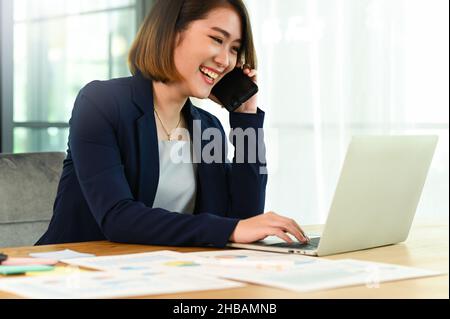 Geschäftsfrau in einem Anzug, die telefoniert und mit einem laptop-lächelnden Gesicht spricht, die junge Frau, die in einem Büro arbeitet und mit f telefoniert Stockfoto