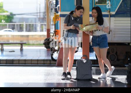 Zwei Mädchen im Teenageralter, die eine Zugfahrt auf dem Bahnsteig für eine Reise planen, ein ausländisches Mädchen im Teenageralter, das einen Laptop mit Kopfhörern um den Hals und ein c hält Stockfoto