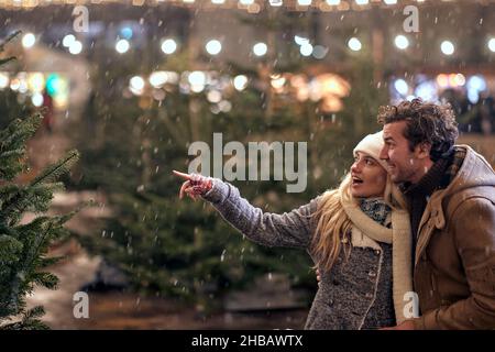 Ein junges verliebtes Paar kommentiert weihnachtsbäume, während es die weihnachtsferien genießt und in einer wunderschönen verschneiten Nacht durch die Stadt läuft. Weihnachten, Neu Stockfoto