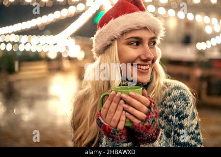 Junge Frau in santa hat heißen Tee auf einem Jahrmarkt in Weihnachten trinken Stockfoto