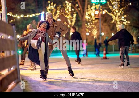 Ein junger Mann hilft seiner Freundin, während er während der weihnachtsferien an einem schönen Abend in der Stadt Schlittschuhlaufen lernt. Christma Stockfoto