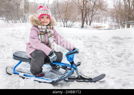 Kleines Mädchen, das im Winter auf einem Schlitten im Park sitzt. Der Winter ist gekommen. Stockfoto