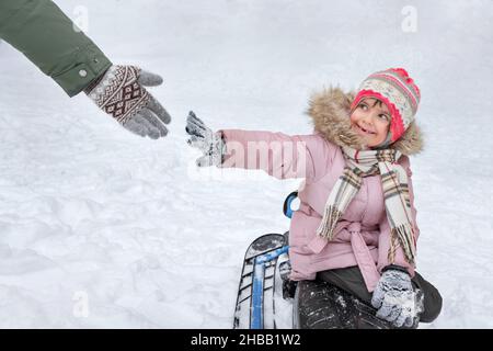 Glückliches zahnloses Mädchen sitzt auf einem Schlitten in einem Winterpark, greift nach der Hand eines Erwachsenen. Stockfoto