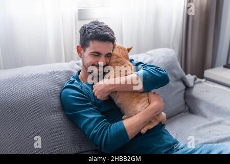 Ein junger Mann, der auf einem grauen Sofa sitzt, umarmt eine braune Katze mit Tabby Stockfoto