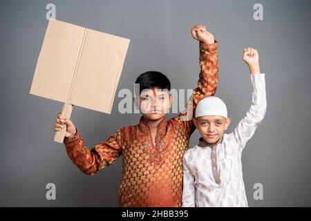 Multiethnische Kinder protestieren mit leerem Schild auf grauem Hintergrund - Konzept des Protests gegen kommunale Gewalt und religiöse Ungleichheit Stockfoto