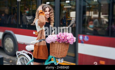 Lockig fröhliche Radfahrerin mit City-Retro-Fahrrad und Blumen im Korbkorb auf dem Hintergrund des roten öffentlichen Busses. Passagiere des öffentlichen Verkehrs in Schutzmasken. Leben in einer Pandemie von Covid-19. Stockfoto