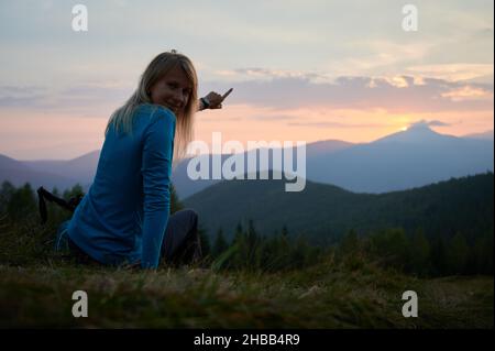 Silhouette eines Mädchens mit einem leichten Lächeln auf dem Gesicht, das auf einem Hügel ruht, das Panorama der Berggipfel genießt und ihre Hand auf die Wolken am Morgenhimmel bei Sonnenaufgang zeigt. Stockfoto