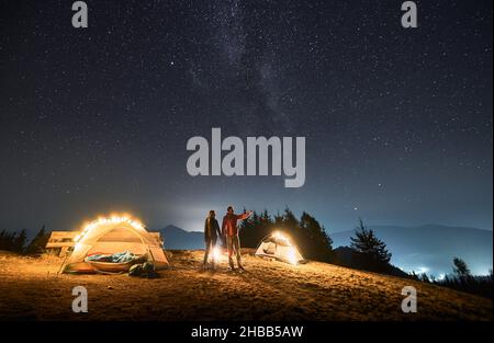 Junge, der in der Nähe von zwei hellen Zelten stand, die Hand seiner Freundin hielt und sie auf die Landschaft vor ihm zeigte. Nächtliches Campen auf einer Bergwiese unter Sternenhimmel. Konzept des Wanderns, Nachtcamping. Stockfoto