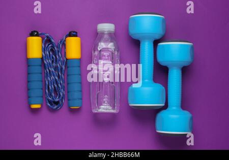 Sportkonzept im flachen Lay-Stil. Kurzhanteln, Springseil, Flasche Wasser. Sportgeräte auf violettem Hintergrund. Draufsicht Stockfoto