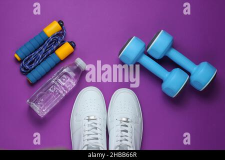 Sportkonzept im flachen Lay-Stil. Kurzhanteln, Turnschuhe, Springseil, Flasche Wasser. Sportgeräte auf violettem Hintergrund. Draufsicht Stockfoto
